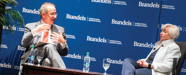 A man talks with a woman while seated in front of a blue Brandeis backdrop